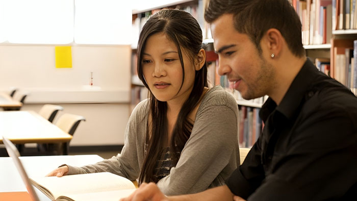 Two students collaborating in a library setting, using a textbook and laptop for academic support.