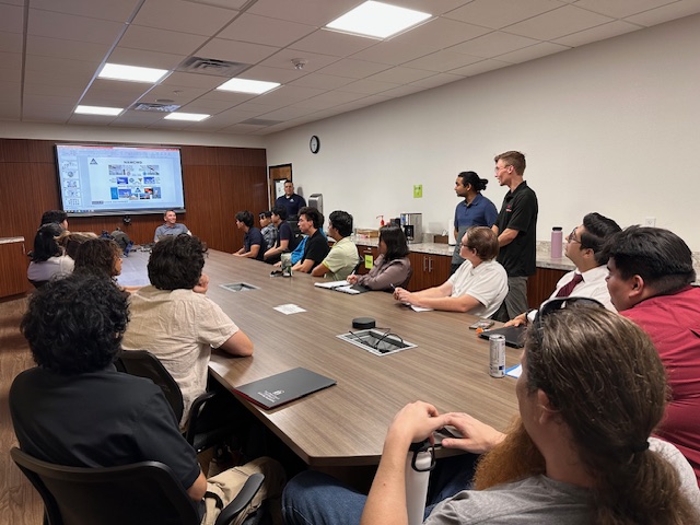 A group of people sits around a conference table to listen to the presenter at the front. 