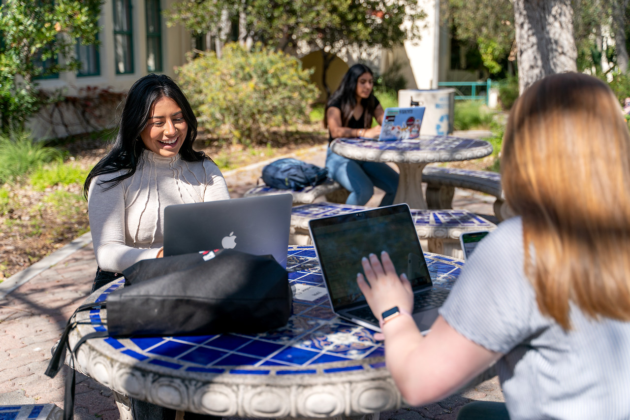 Two female students sit at a lunch table with their laptops and laughing.