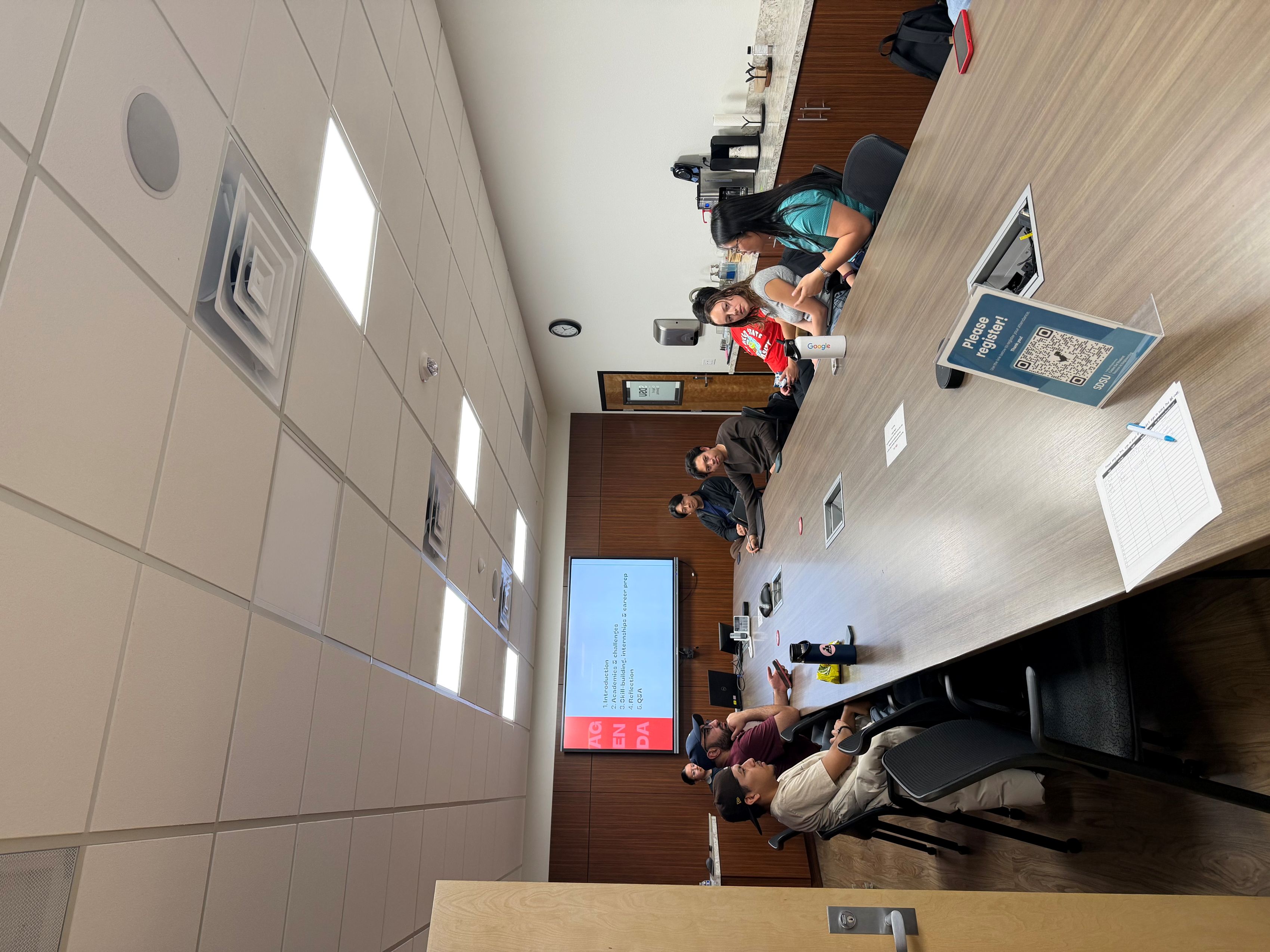A group of people sits at a conference table to discuss. The projector shows the agenda for a student panel.